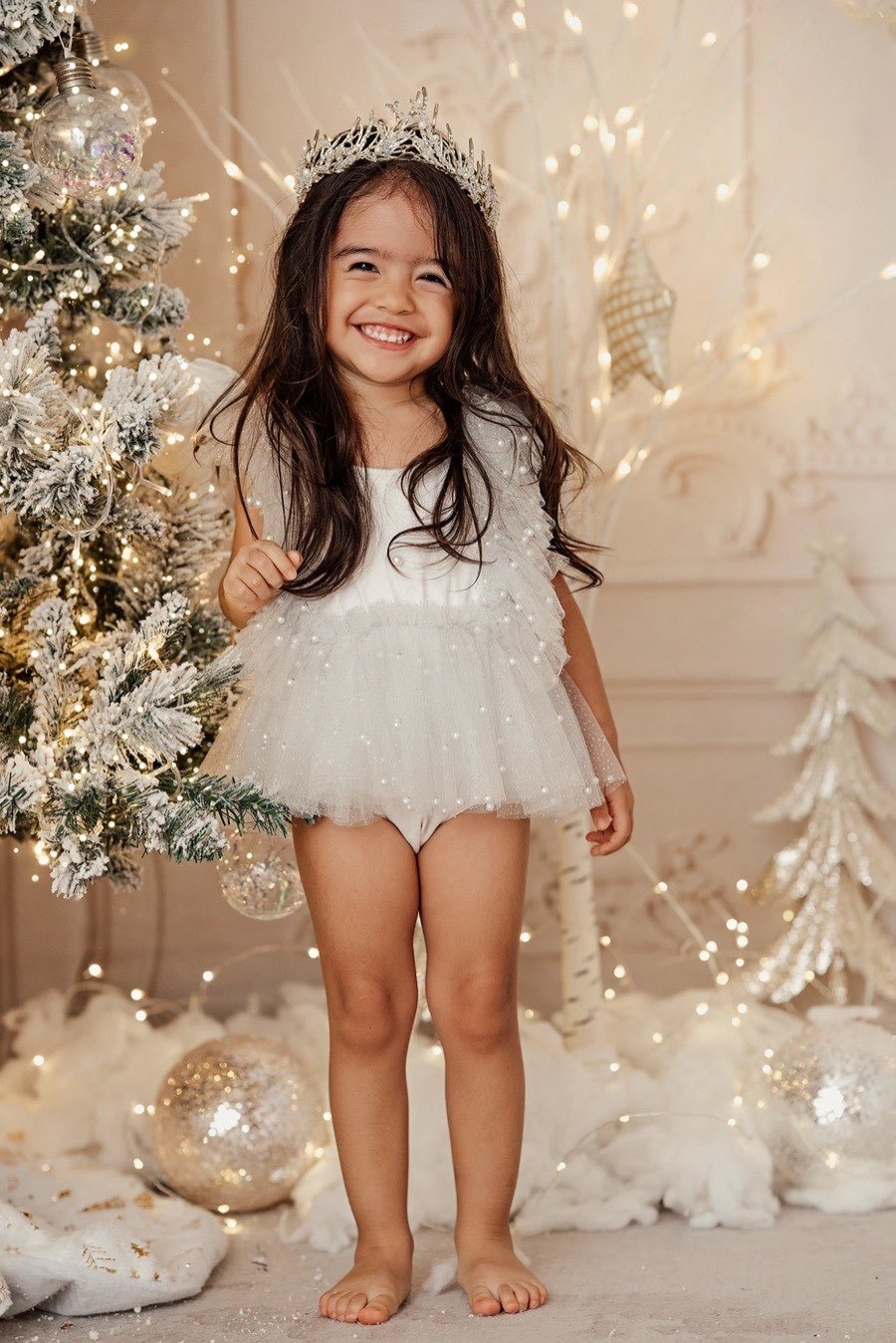Niña con corona y vestido de tul junto al árbol de Navidad nevado